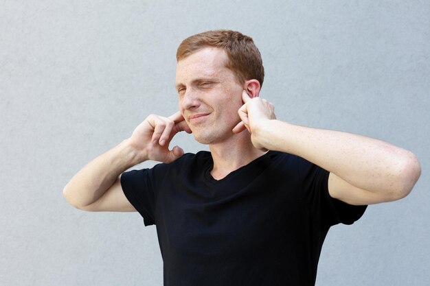 fashion style emotions and people concept Close up portrait of a redhead of a beautiful manly guy with freckles frowning his face closing ears with palms annoyed and tired of noise