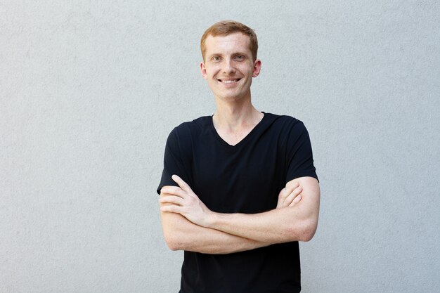 Fashion, style, emotions and people concept - Close up portrait of a redhead of a beautiful manly guy with freckles on a gray background black T-shirt. crossed hands on chest