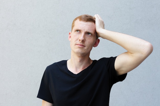 Fashion, style, emotions and people concept - Close up portrait of a redhead of a beautiful manly guy with freckles on a gray background black T-shirt. frightened expression trying to remember