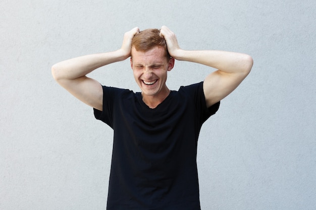 Fashion, style, emotions and people concept - Close up portrait of a redhead of a beautiful manly guy with freckles on a gray background black T-shirt. Holds his hands at the head and laughs