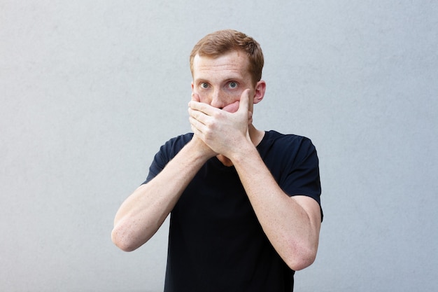 Fashion, style, emotions and people concept - Close up portrait of a redhead of a beautiful manly guy with freckles on a gray background. covers his mouth with his hands so as not to tell a secret