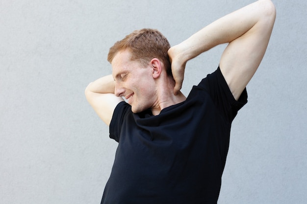 Fashion, style, emotions and people concept - Close up portrait of a redhead of a beautiful manly guy with freckles on a gray background . Holds his hands behind his neck and smile, stretches