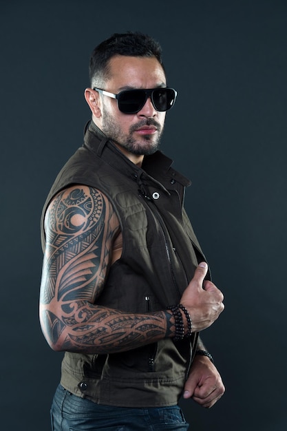 Fashion style and trend Tattooed man with biceps and triceps Tattoo model with beard on unshaven face Bearded man with tattoo on strong arms Fashion macho in trendy sunglasses