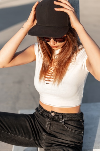 Fashion swag woman in american black stylish cap in sunglasses in summer casual clothes sits on outdoor parking in sunny day in city. Youth modern style