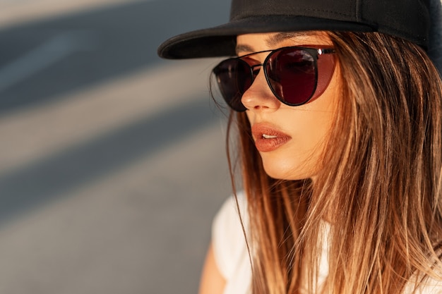 Fashion swag young girl with pretty lips in tender skin in stylish black baseball cap and trendy sunglasses on street at amazing orange sunset. Cool modern woman on sun. Close-up portrait.