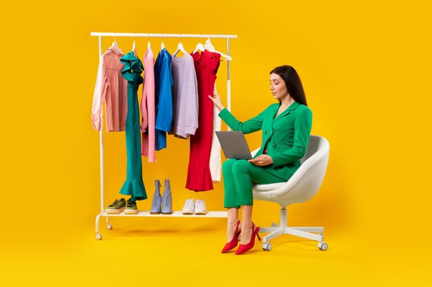 Fashion trends beautiful woman stylist sitting near lot of clothes on garment rack and using laptop