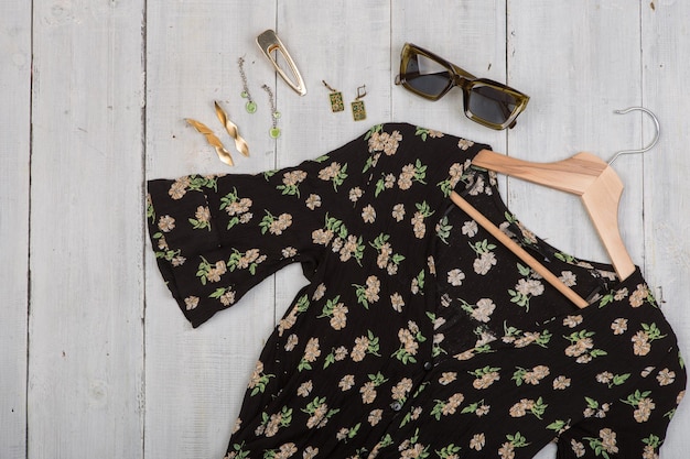 Fashion trends - sunglasses, black dress in floral print on hanger and jewelry: barrette, silver and gold earrings on white wooden desk