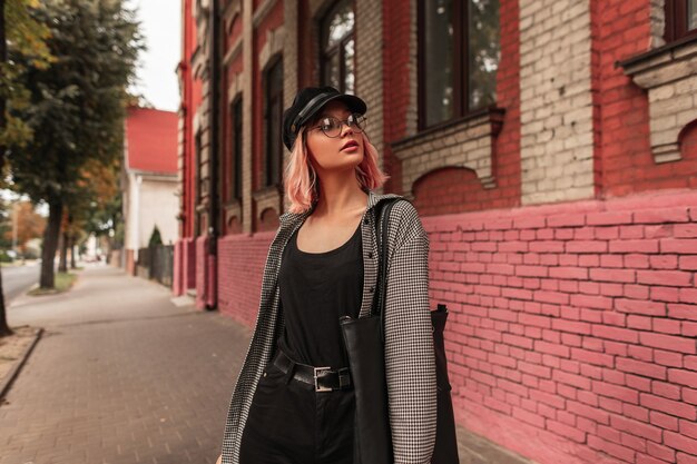 Fashion urban beautiful girl with pink hair in fashion clothes, a shirt and jeans with glasses and a vintage cap is walking on the street near a grunge brick building