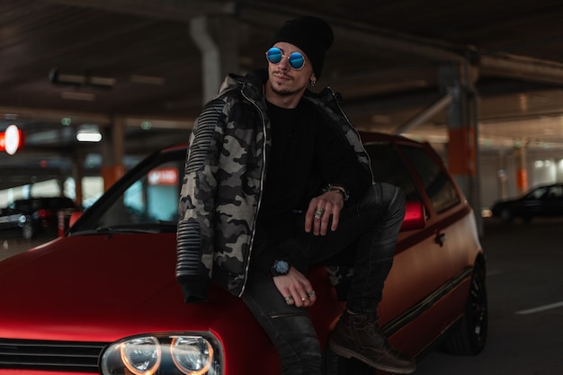 Fashion urban man model with sunglasses and hat in stylish winter military jacket and pullover stands near a red car at parking lot