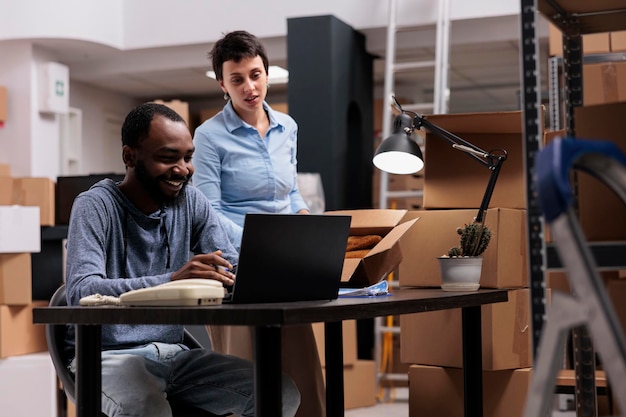 Fashion warehouse manager putting trendy blouse in carton box\
while supervisor man analyzing shipment on laptop computer before\
delivering order. team working at transportation logistics in\
storehouse