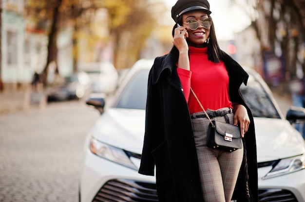 Fashion woman in coat, newsboy cap and sunglasses posed at street against white business car.