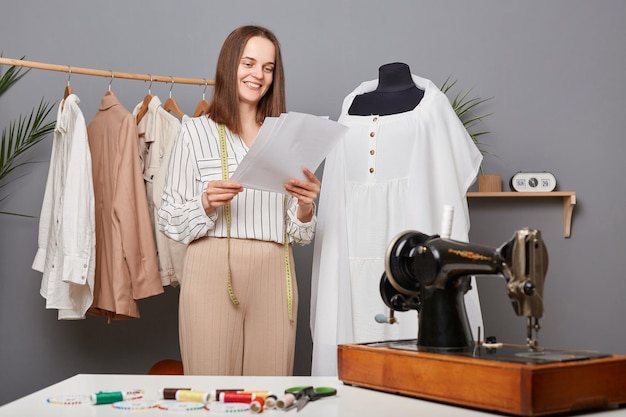 Модная женщина-дизайнер, держащая бумаги с эскизом в мастерской, думает о новой коллекции, улыбающаяся швея, создающая новые наряды