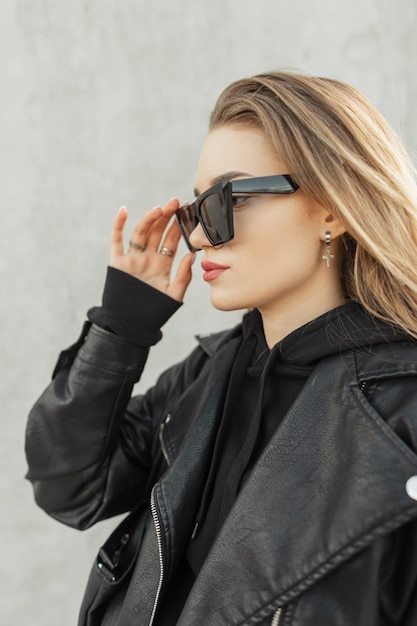 Fashion woman hipster with trendy leather black jacket with black stylish hoodie puts on a cool sunglasses on a gray concrete background Urban female rock style clothes