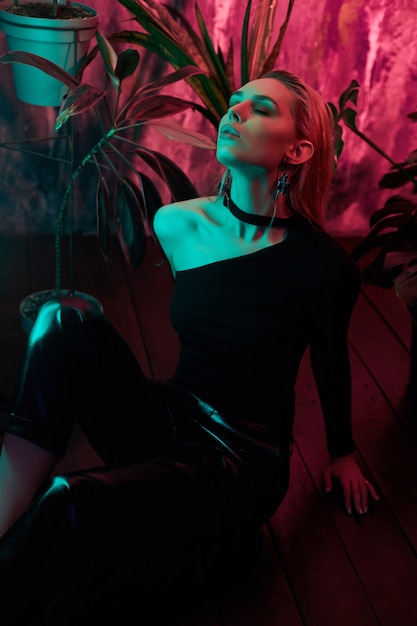 Fashion woman sitting on the floor in tropical foliage neon light. Wet hair, perfect figure and makeup