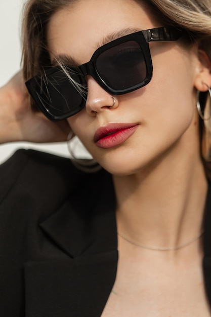 Fashion women's cool summer street portrait of a beautiful stylish young woman with a nose piercing with black fashion sunglasses wearing black elegant outfit on a sunny day