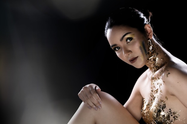 Fashion Young Asian Woman eyes black wrapped hair beautiful make up fashion decorate with Golden Foil or Gold leaf all over chest. Studio Lighting dark Background