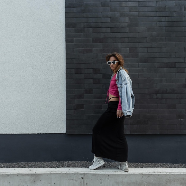 Fashion young beautiful woman model with trendy white sunglasses in fashionable denim clothes with jeans jacket pink top and black long skirt with sneakers walks and poses near a modern wall