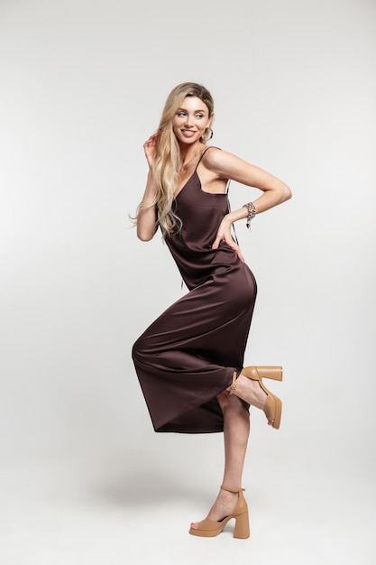 Fashion young elegance beautiful blonde happy woman with smile in stylish long strappy brown dress and shoes stands and poses on a white background in studio Female beauty and fashion style