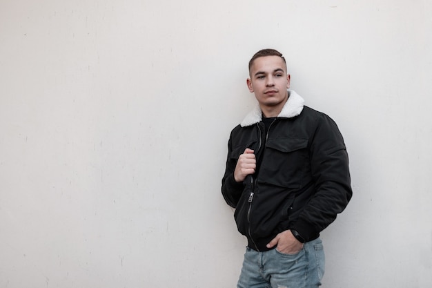 Fashion young handsome man in stylish black outwear stands near gray wall