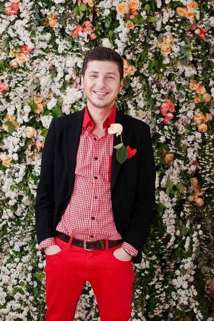 Фасонируйте молодого человека в черном костюме и красных брюках вскользь на стене декора предпосылки флористической.