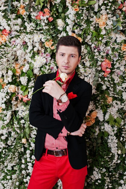 Fashion young man with rose at hand, in black suit and red pants on background floral decor wall. 