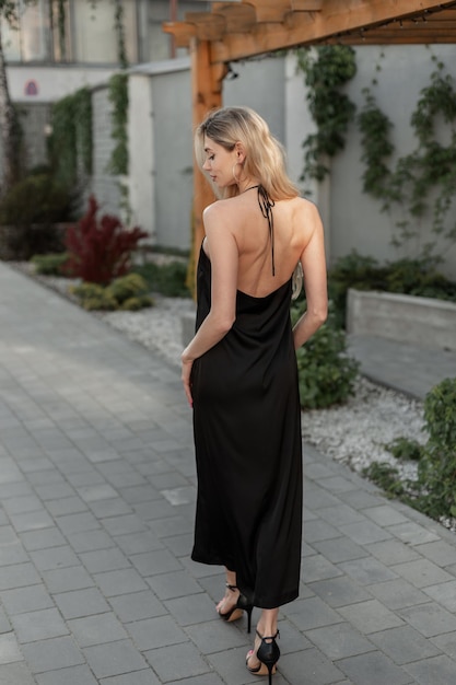 Fashion young pretty woman blonde Caucasian model in elegant black fashion open back strappy long dress with heels shoes walks in the city Urban female evening style and beauty