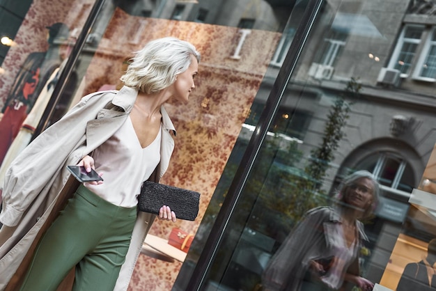 Foto moda giovane donna alla moda che cammina per la strada della città che tiene smartphone guardando la riflessione a