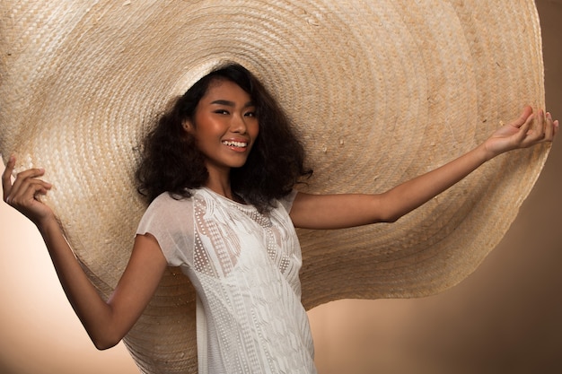 Fashion Young thin Asian Woman Tan skin eyes beautiful make up fashion white dress posing attractive glam look very big straw hat. Studio Lighting beige yellow cream Background, summer beach hot sun