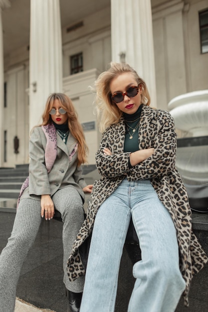 Fashion young trendy woman with girlfriends in stylish clothes with sunglasses leopard coat and classic blue jeans sits and poses in the city near vintage columns