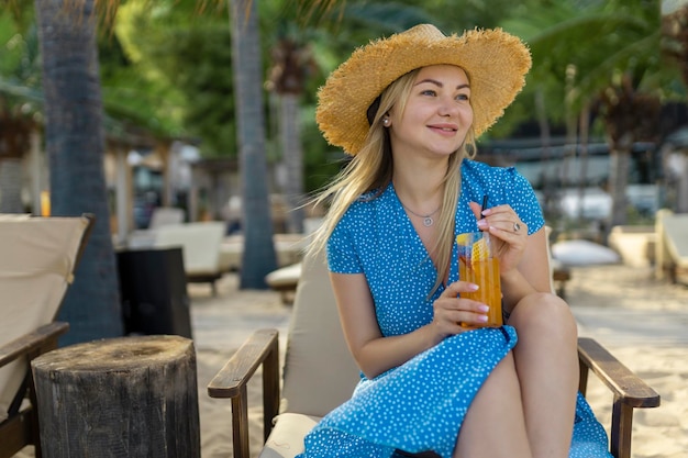 Модная молодая женщина пьет коктейль в пляжном баре Веселая привлекательная блондинка в платье улыбается и пьет коктейль на открытом воздухе Счастливая женщина наслаждается летним временем на пляже