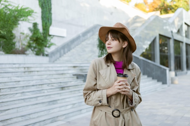 フェルトの帽子と秋のトレンチコートを着たファッションの若い女性が都市公園でコーヒーを飲みます。