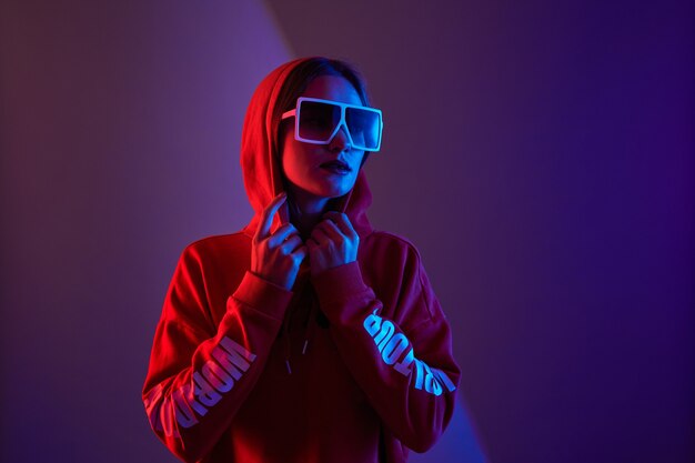 Fashion young woman model wear hoodie and eyewear sunglasses in neon light studio background.