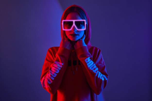 Fashion young woman model wear hoodie and eyewear sunglasses in neon light wall. Close up.