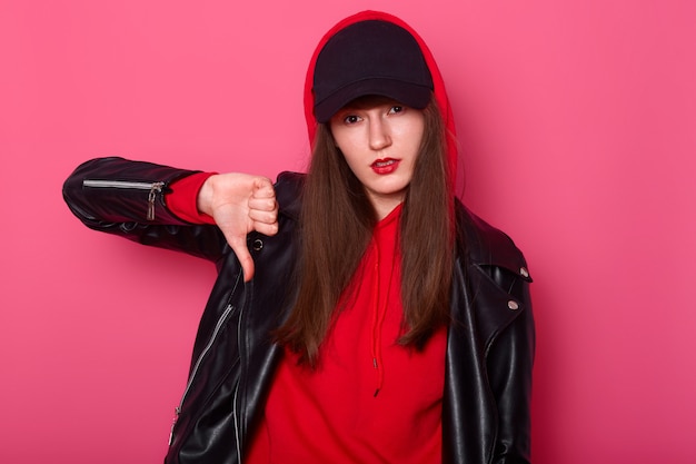 Fashion young woman uses bright red lipstick, wears stylish shirt, leather jacket and black cap. Cheeky tenager shows finger down sign