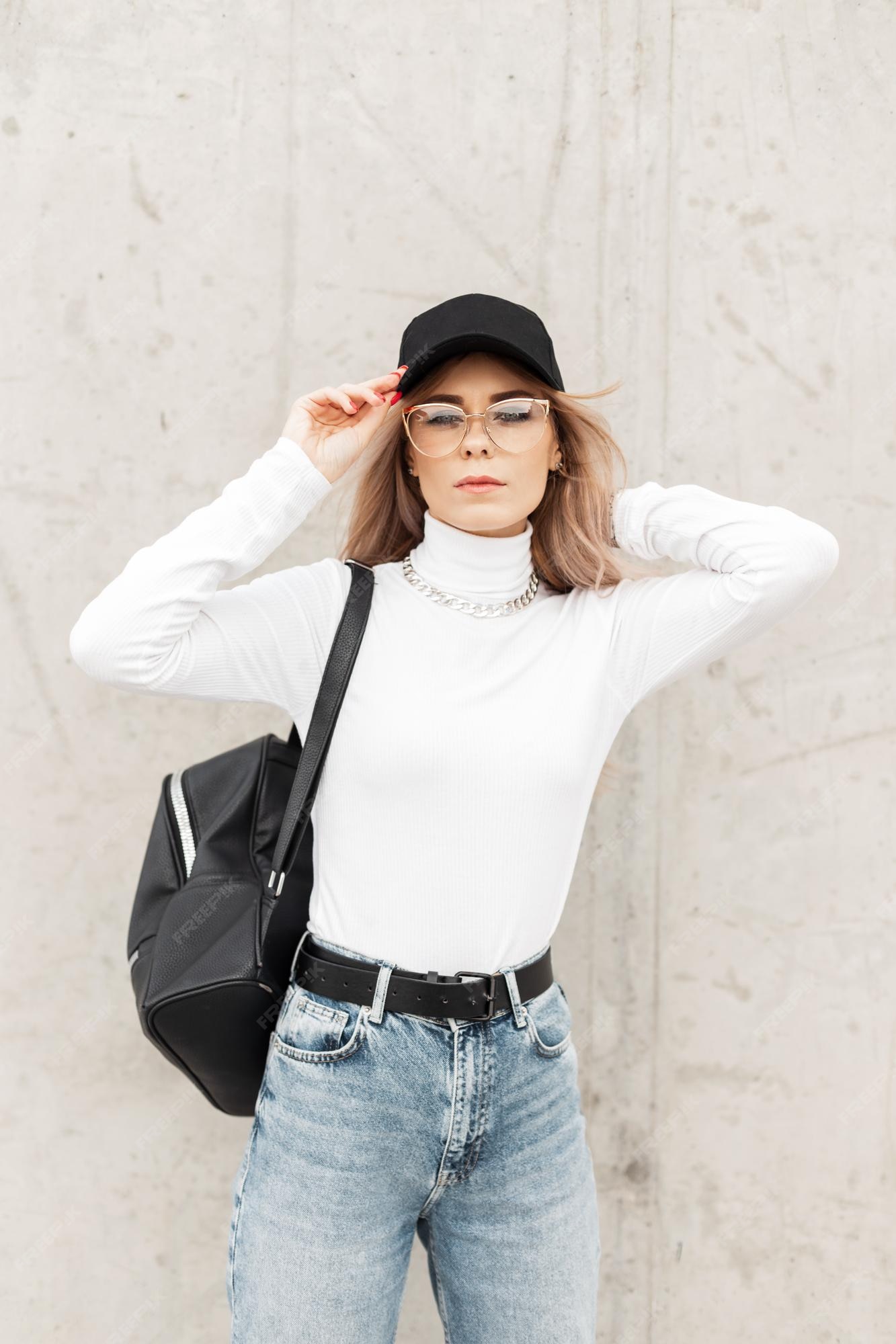 Premium Photo | Fashion young woman with glasses in casual trendy clothes  with black cap white sweatshirt jeans and bag stands near a gray concrete  wall