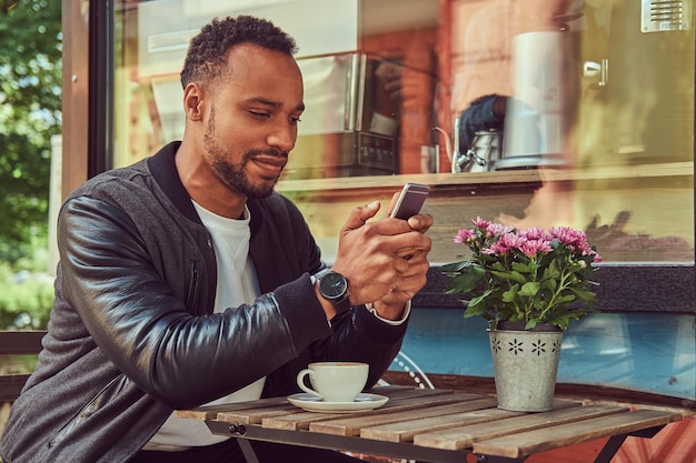 Fashionable african-american bearded male sitting near a coffee\
shop with a cup of coffee, using a smartphone.