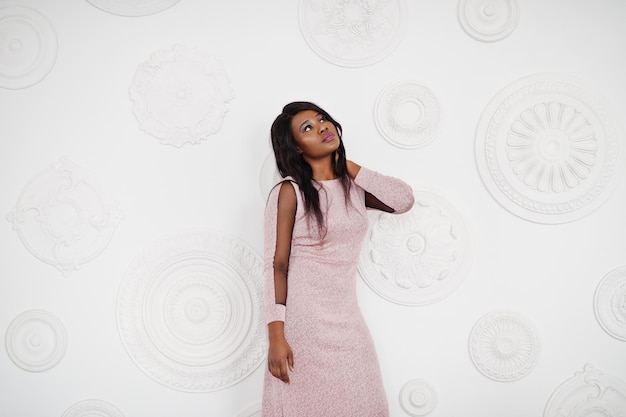 Fashionable african american model woman in pink brilliant evening dress posed against white decorative wall