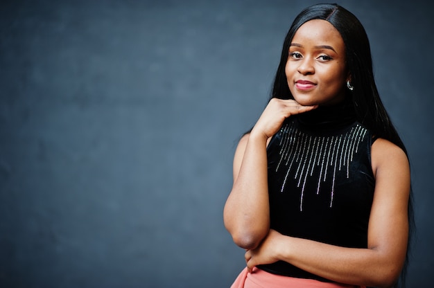 Fashionable african american woman in peach pants and black blouse pose against gray wall.