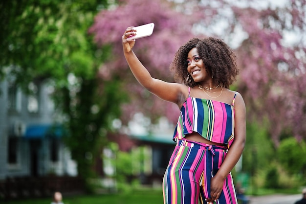 Donna afroamericana alla moda in tuta a righe rosa posata in strada fiorita primaverile e facendo selfie con il telefono cellulare