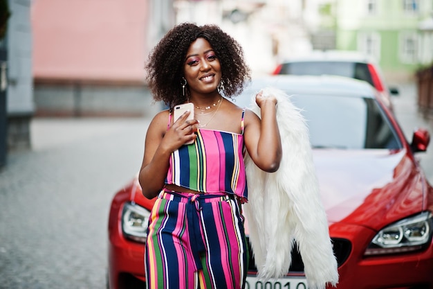 Fashionable african american woman in pink striped jumpsuit with fluffy faux fur coat posed against rich red luxury car with mobile phone at hand