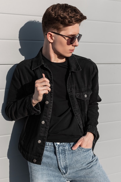 Модный американский молодой человек в винтажных солнцезащитных очках в молодежной стильной повседневной джинсовой одежде