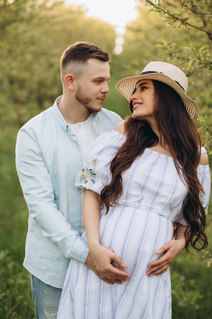 Фото Модная и стильная счастливая беременная женщина и ее муж одеты в пастельные бело-голубые тона в саду на закате