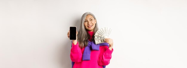 Foto una donna anziana asiatica alla moda che mostra soldi, dollari e uno schermo vuoto dello smartphone dimostra una persona online