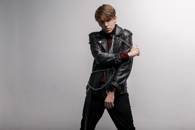 fashionable attractive man in a black leather jacket in retro style in red stylish golf and black pants posing in the afternoon in a room near the white wall. cute stylish guy