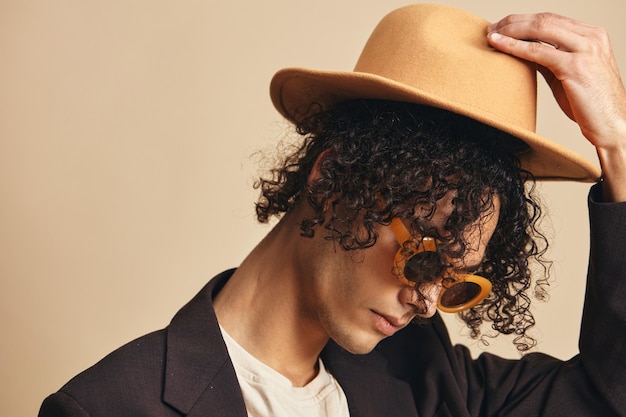 Fashionable awesome tanned curly man in brown jacket trendy sunglasses wear classic hat posing isolated on beige pastel background Fashion New Collection offer Retro style concept Free place for ad