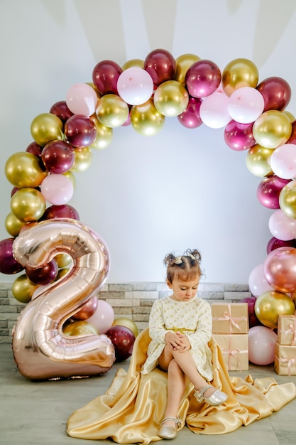 Fashionable baby girl in a white clothing celebrating her second birthday posing for camera and looking down. Lovely baby girl birthday photoshoot