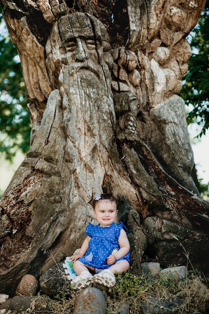 Fashionable baby with blue dress sitting at old giant tree and smiling