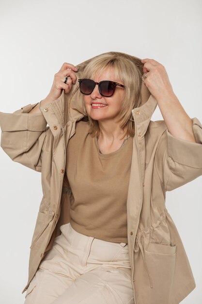 Fashionable beautiful aged woman with vintage sunglasses and a stylish beige jacket puts on a hood on a white background in the studio