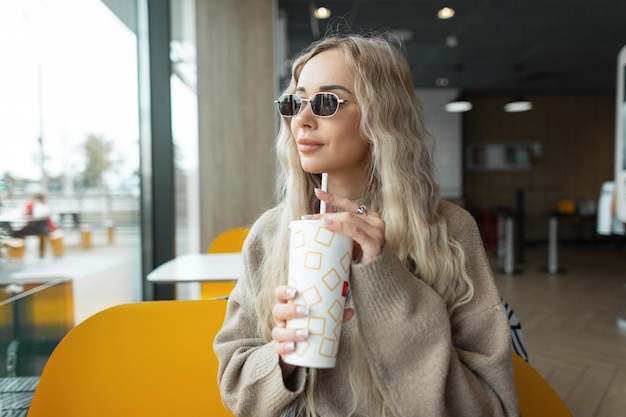 Fashionable beautiful blond woman hipster sits in a cafe holds a paper cup of cola and drinks drink