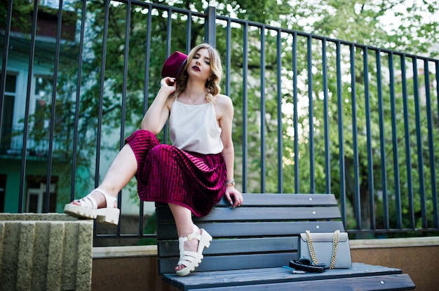 Fashionable and beautiful blonde model girl in stylish red velvet velour skirt, white blouse,hat and handbag, posed on bench.